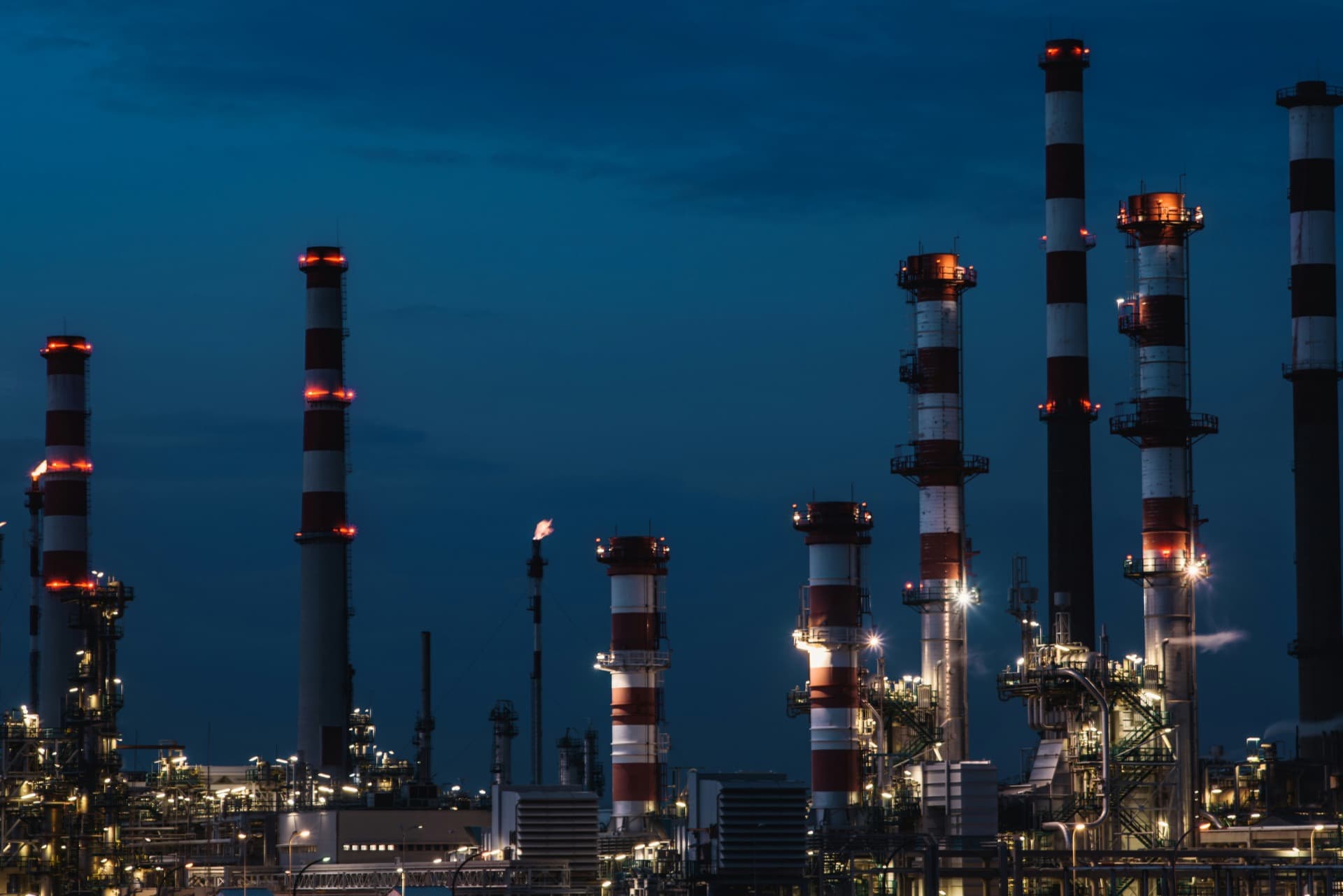 Industrial refinery smokestacks illuminated against a deep blue dusk sky